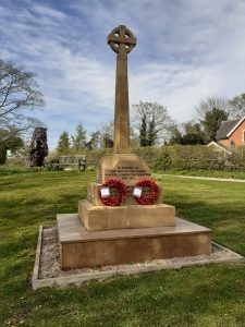 Photograph of a War Memorial. Stone obelisk with two red poppy remembrance day wreaths laid at its base.